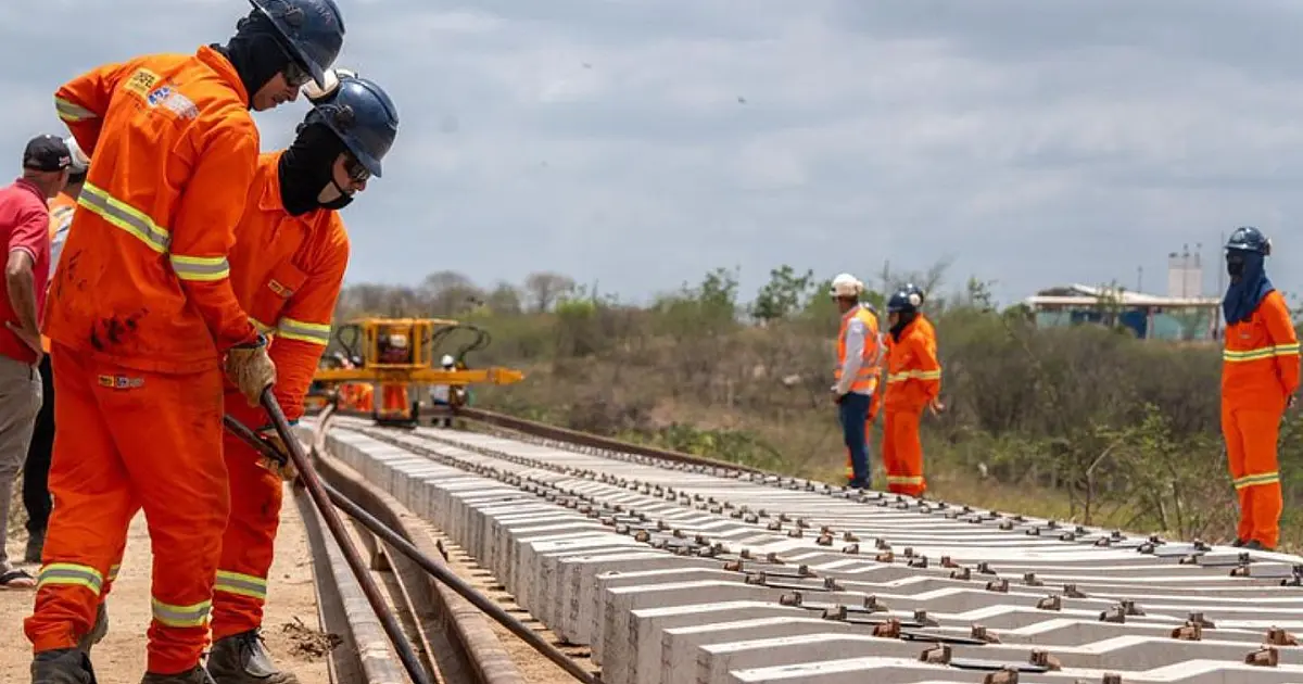 Trecho de ferrovia em construção com trilhos, máquinas e trabalhadores em área aberta no Nordeste.