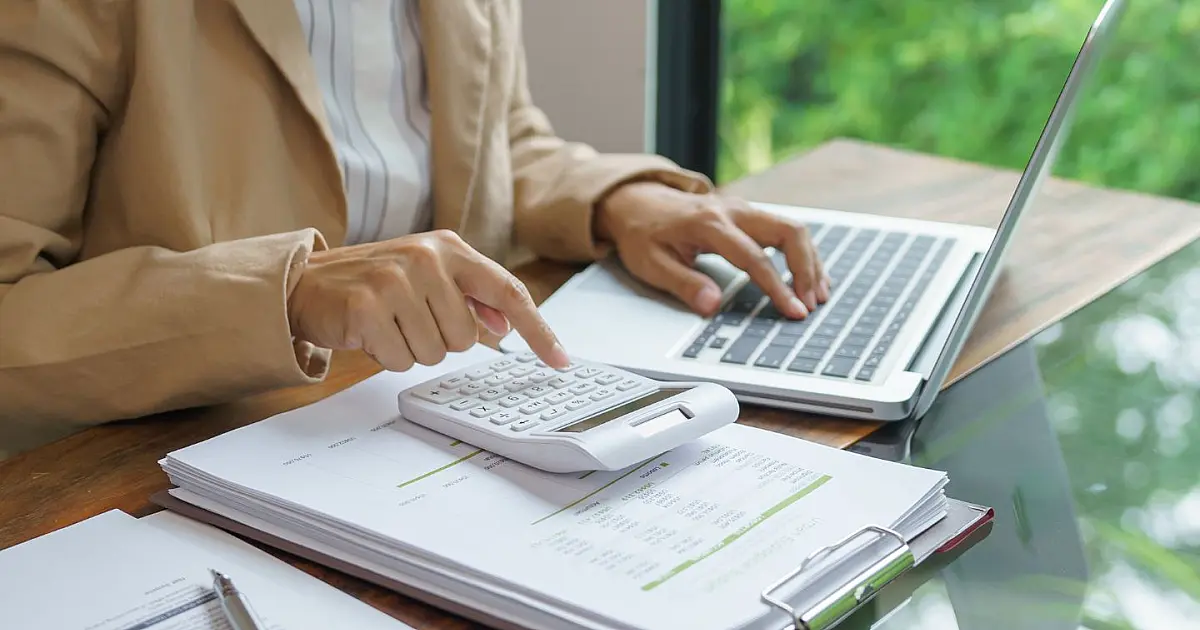 Pessoa utilizando calculadora e computador com documentos fiscais espalhados sobre a mesa.