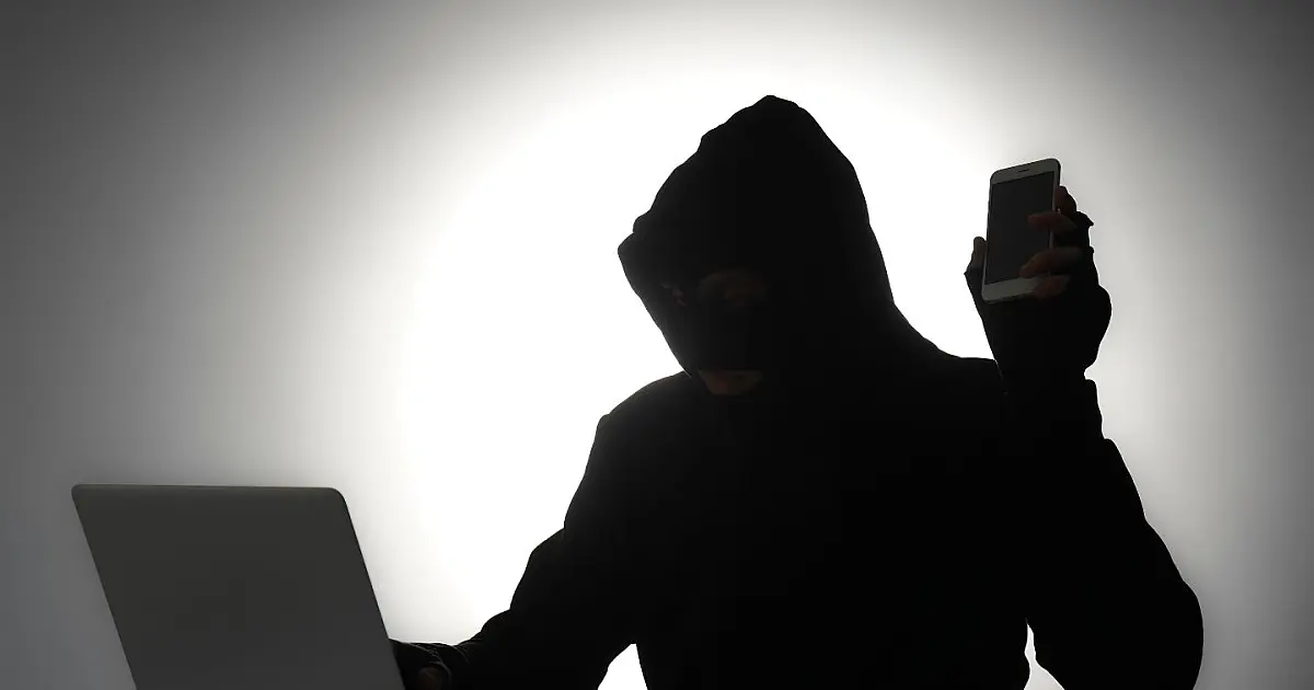 Silhueta de homem usando celular e notebook em ambiente escuro, representando fraude digital.