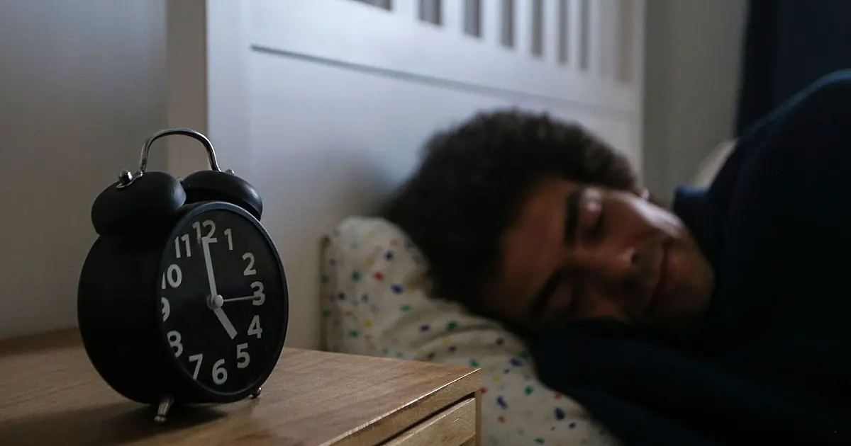 Homem dormindo ao lado de relógio sobre mesa de cabeceira durante a noite.