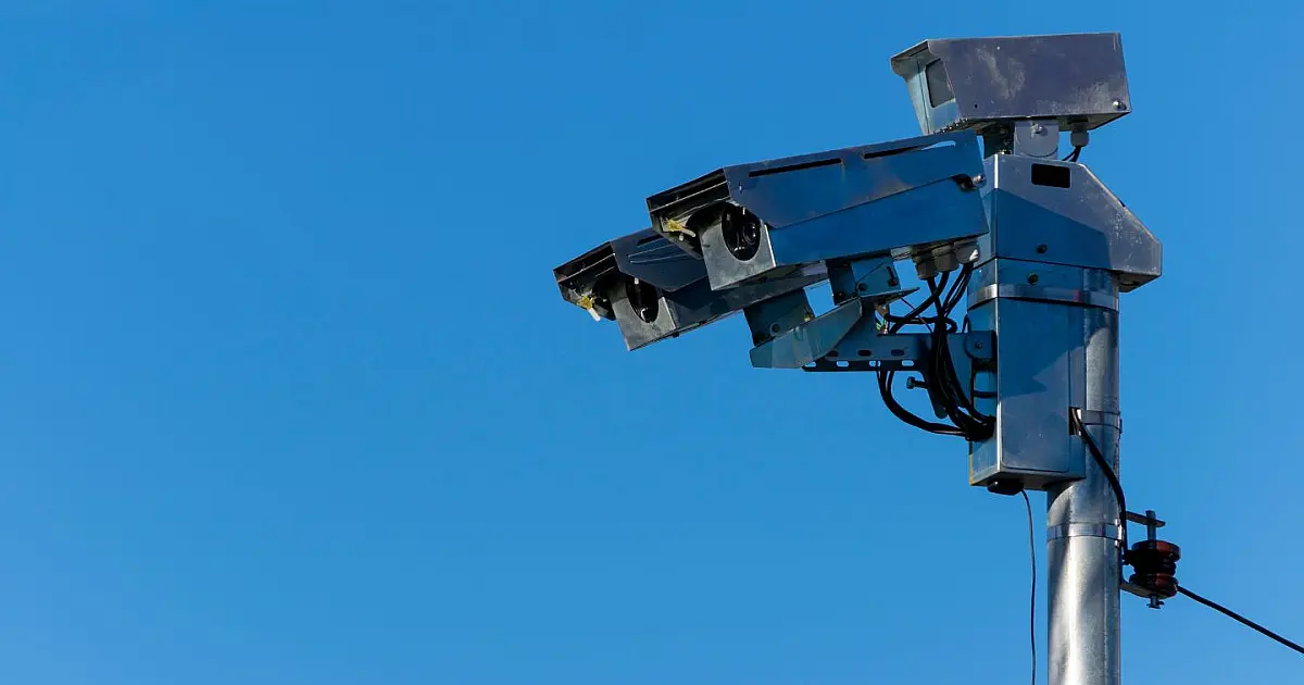Detalhe de um radar fotográfico cinza de controle de velocidade instalado no topo de um poste metálico à margem de uma avenida.