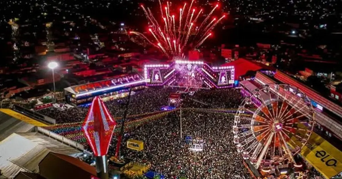 Fotografia aérea noturna mostrando o Pátio de Eventos de Caruaru completamente lotado de pessoas, iluminado por luzes coloridas do palco principal e cercado pela arquitetura urbana da cidade.