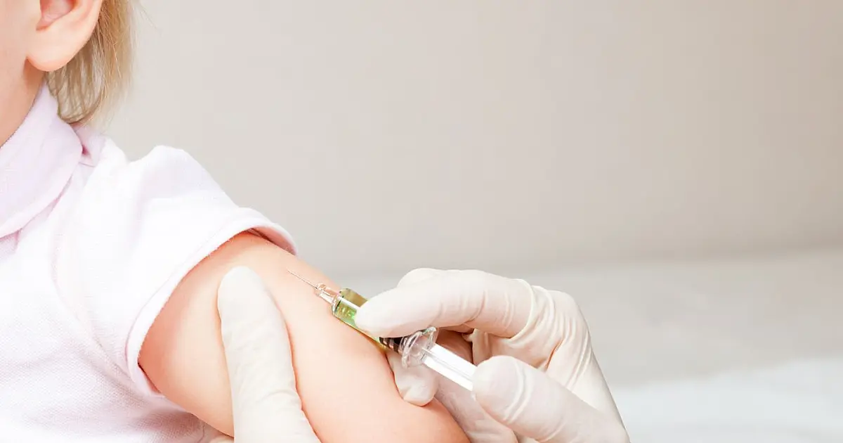 Close em uma profissional de saúde usando luvas aplicando uma vacina no braço de uma criança, que está sentada no colo de um adulto.