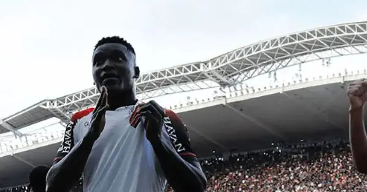 Jogador de futebol com camisa listrada em vermelho e preto comemorando um gol de braços abertos no gramado.