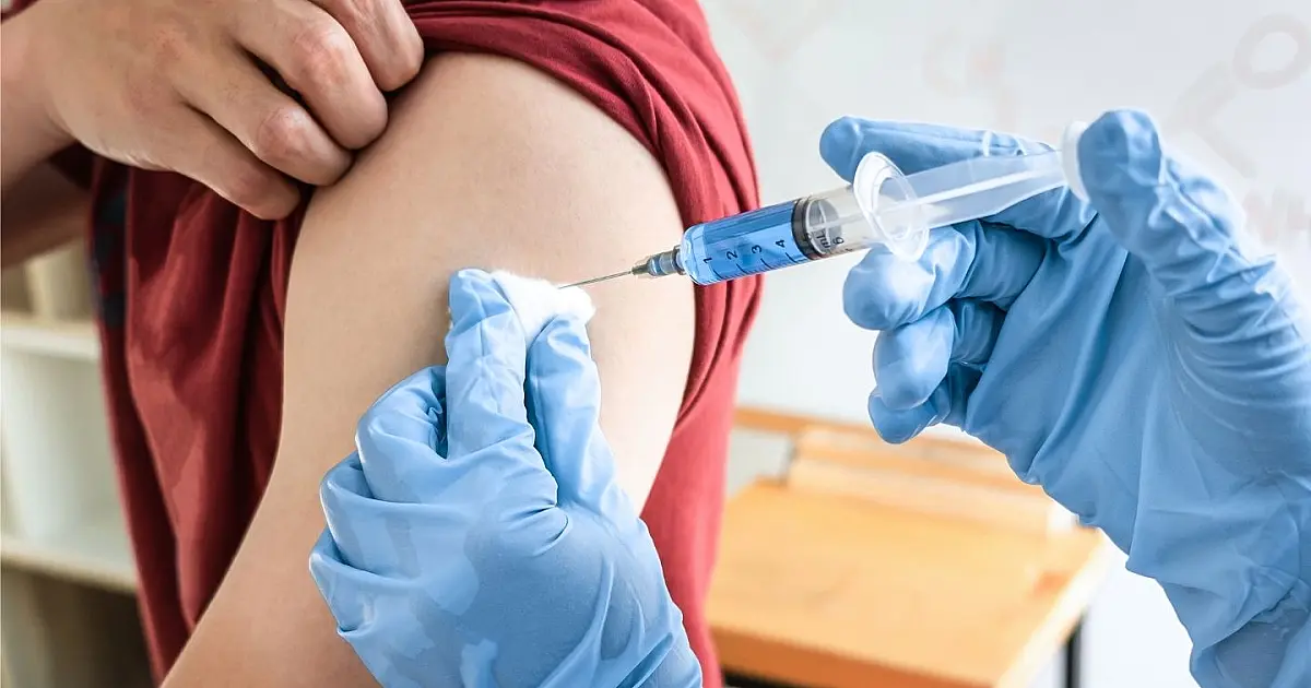 Profissional de saúde utilizando luvas e aplicando uma dose de vacina com seringa no braço de um paciente.
