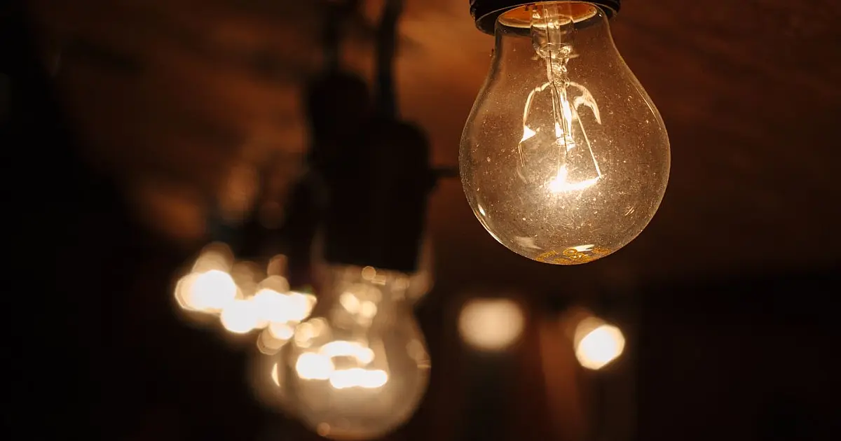 Uma lâmpada de vidro tradicional com filamento interno acesa emitindo uma luz amarelada e quente contra um fundo escuro.