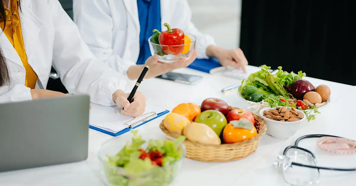 Uma nutricionista de jaleco conversando e orientando um paciente em um consultório, com uma cesta de frutas frescas sobre a mesa entre eles.