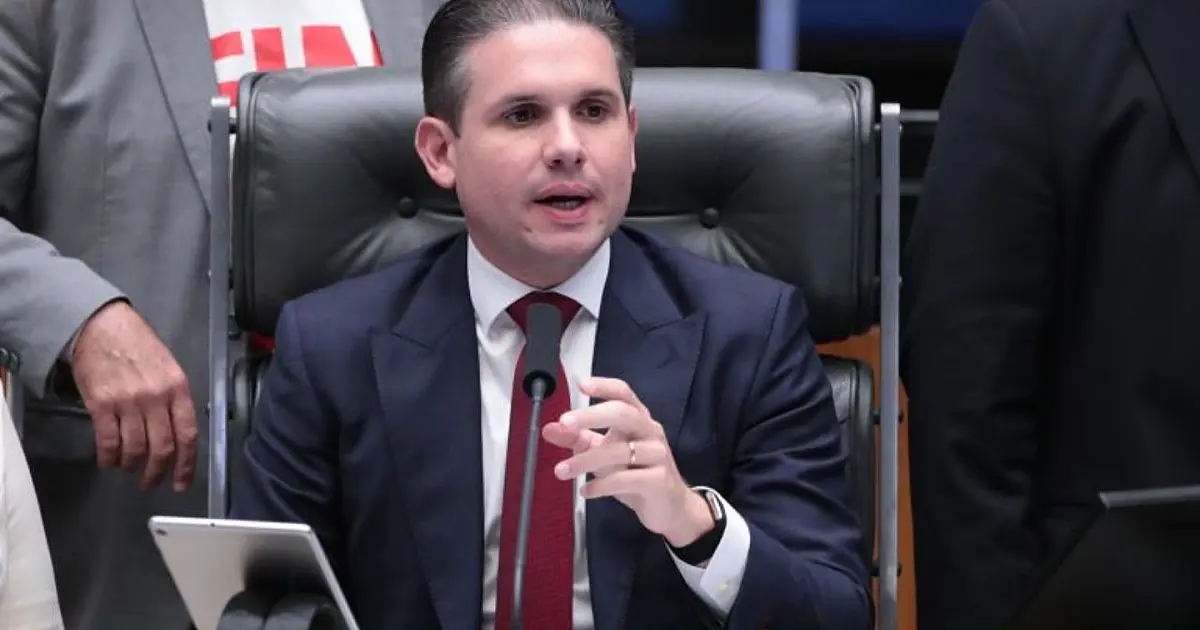Retrato em plano médio do deputado Hugo Motta vestindo terno escuro e gravata, falando formalmente em um ambiente institucional com microfones à frente.
