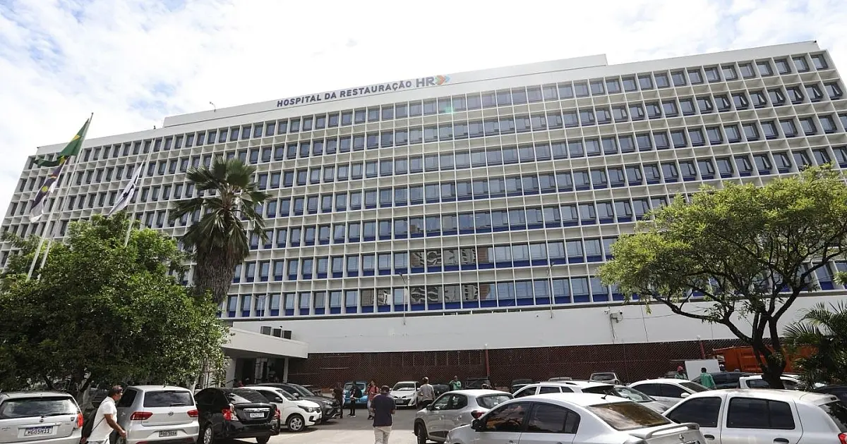 Visão externa da fachada principal de um grande prédio hospitalar público com letreiro institucional visível.