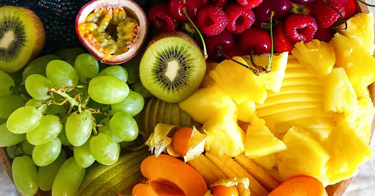 Vista superior de uma travessa de madeira decorada com frutas tropicais e exóticas coloridas e cortadas, com destaque para pequenos frutos arredondados e escuros em primeiro plano.