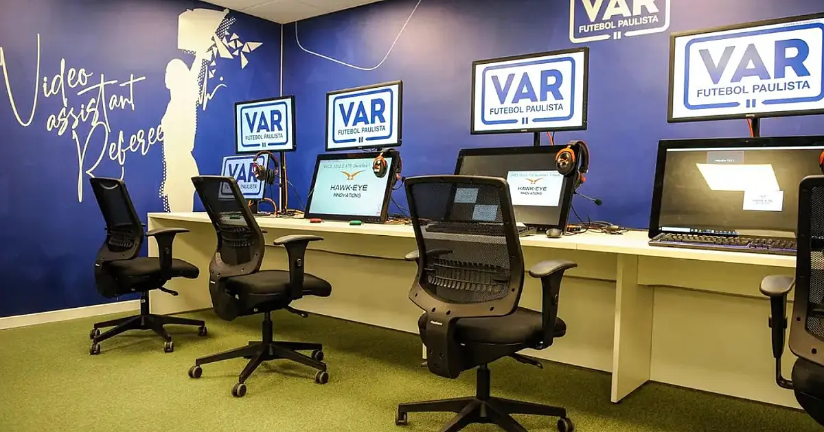 Vista de uma sala tecnológica interna com diversos monitores de alta definição exibindo replays de lances de futebol sob diferentes ângulos de câmera, com operadores sentados em cadeiras de escritório.