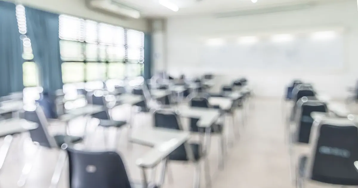 Interior de uma sala de aula de cursinho vazia e levemente desfocada, com várias fileiras de carteiras escolares de madeira voltadas para um quadro branco ao fundo.