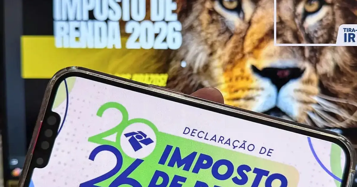 Dedos de uma pessoa digitando informações financeiras no teclado de um computador com tela exibindo planilhas desfocadas ao fundo.