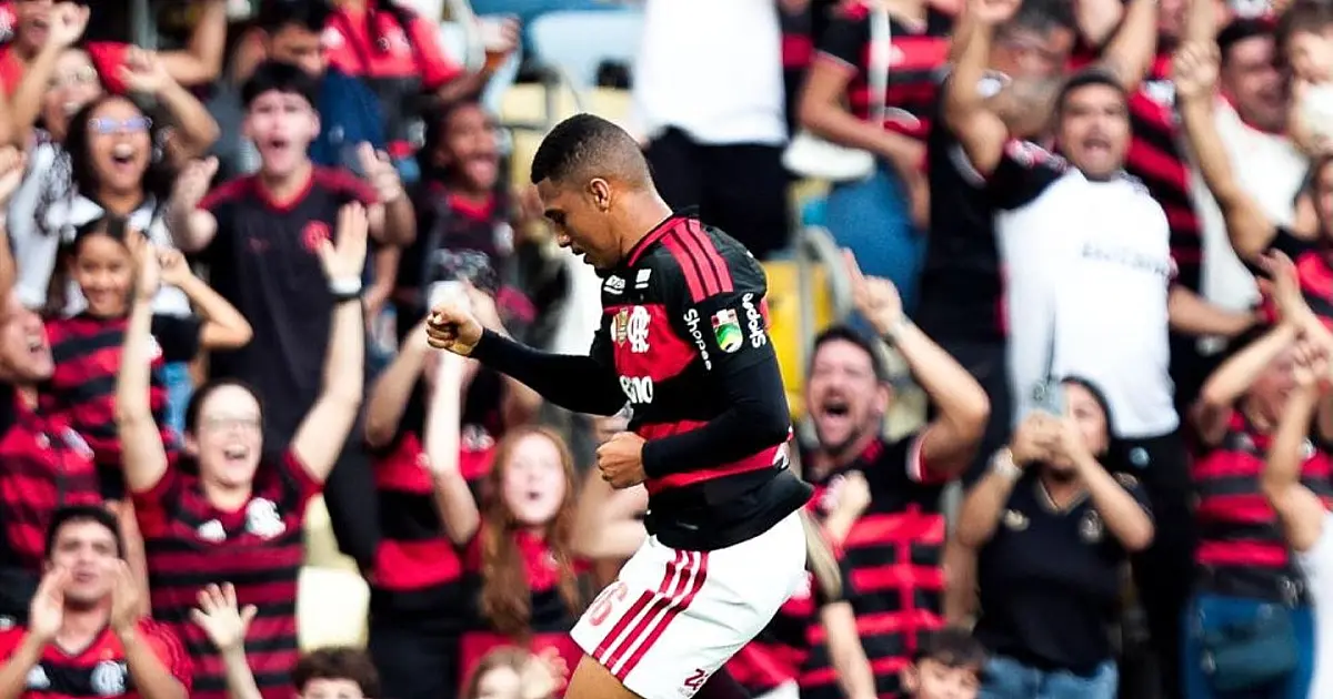 Fotografia em plano médio focando o atacante Samuel Lino sorrindo e correndo pelo gramado com os braços abertos em comemoração, vestindo a camisa listrada em vermelho e preto do Flamengo.