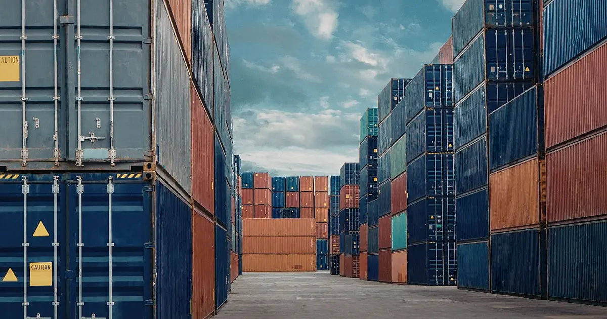 Imagem em plano aberto mostrando um pátio portuário repleto de contêineres coloridos empilhados, com grandes guindastes ao fundo operando sob um céu nublado.