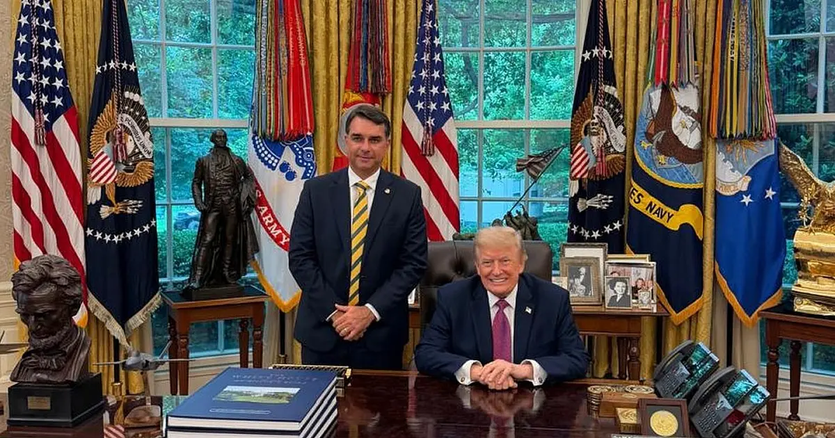 Fotografia em plano médio do senador Flávio Bolsonaro apertando a mão do presidente Donald Trump, ambos sorrindo, no interior decorado do Salão Oval da Casa Branca.
