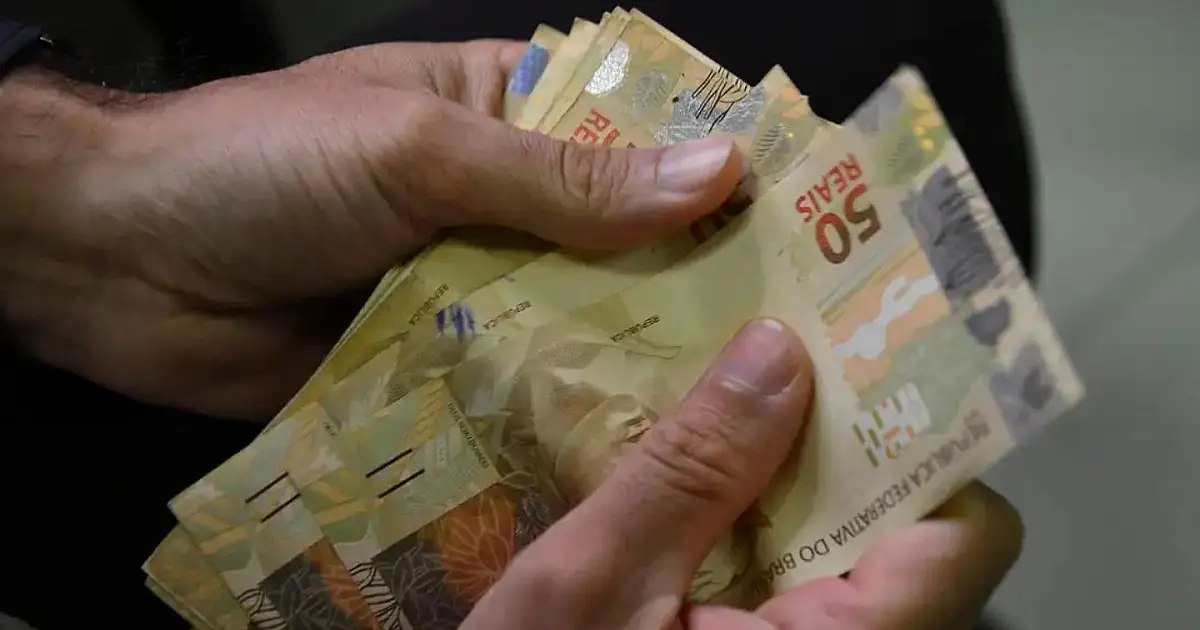 Mãos de uma pessoa manuseando várias cédulas de dinheiro em papel da moeda nacional sobre uma mesa de madeira.
