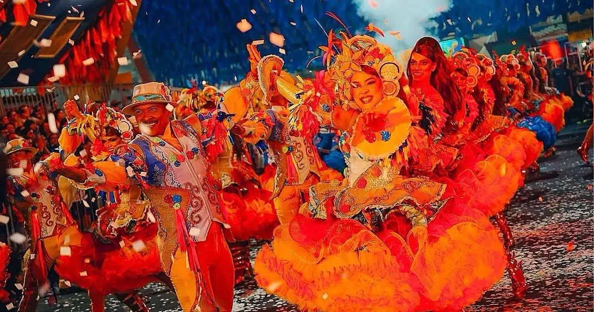 Casais de quadrilha junina com trajes coloridos e tradicionais dançando de forma sincronizada em um grande salão decorado com bandeirinhas.