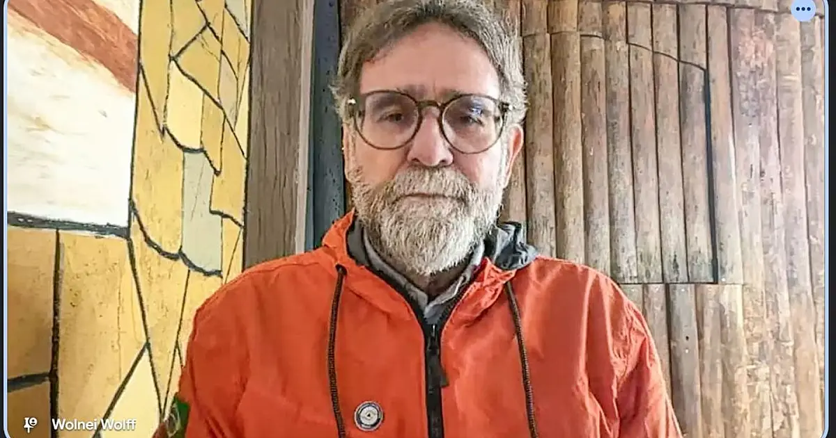 Captura de tela de uma transmissão de vídeo ao vivo mostrando o secretário da Defesa Civil prestando declarações em uma sala de escritório formal