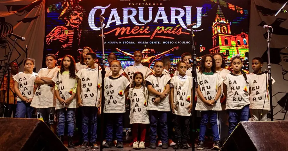 Grupo de jovens vestidos com roupas coloridas típicas de quadrilha junina, dançando e sorrindo no palco sob iluminação cênica de um festival de São João à noite