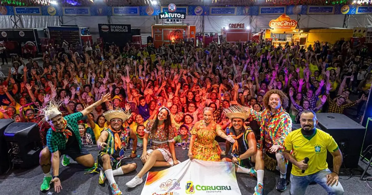 Grande grupo de pessoas sorridentes dançando de braços levantados em uma praça pública decorada com bandeirolas juninas coloridas durante a noite