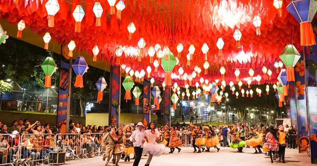 Grupo de jovens estudantes caracterizados com trajes juninos coloridos dançando quadrilha em uma arena iluminada