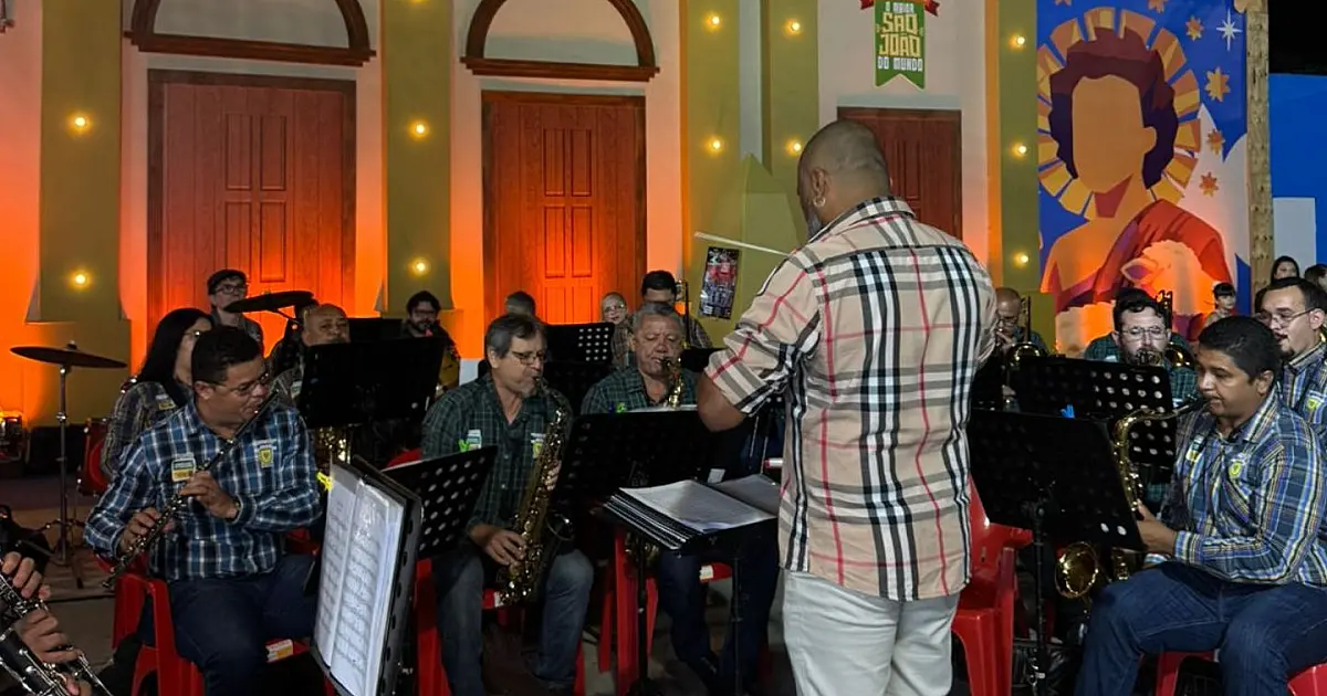 Músicos de uma filarmônica fardados com uniformes formais tocando instrumentos de sopro de metal, como trompetes e trombones, sob a iluminação de um palco