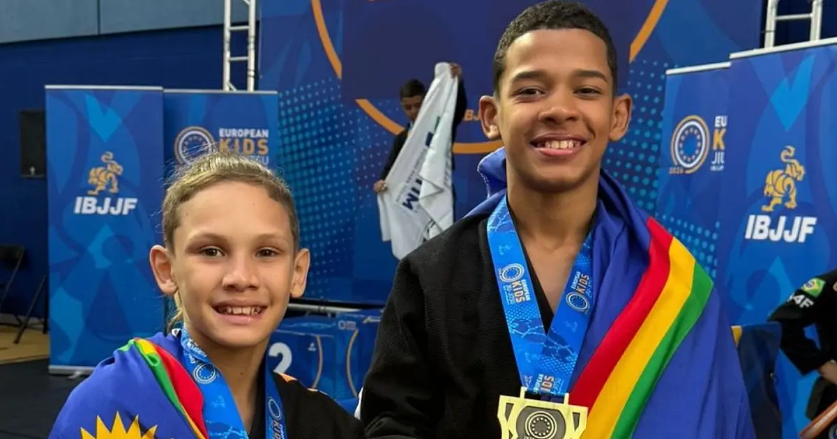 Jovens esportistas sorridentes vestindo agasalhos oficiais com as cores da bandeira de Pernambuco, posando orgulhosamente com medalhas penduradas no pescoço em um ambiente de aeroporto.