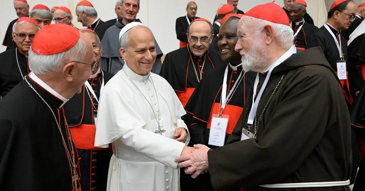 Vista ampla do Papa Leão XIV vestindo batina branca tradicional, sentado ao centro de uma longa mesa diretiva na Sala Paulo VI, cercado por dezenas de cardeais vestindo batinas pretas com detalhes em vermelho