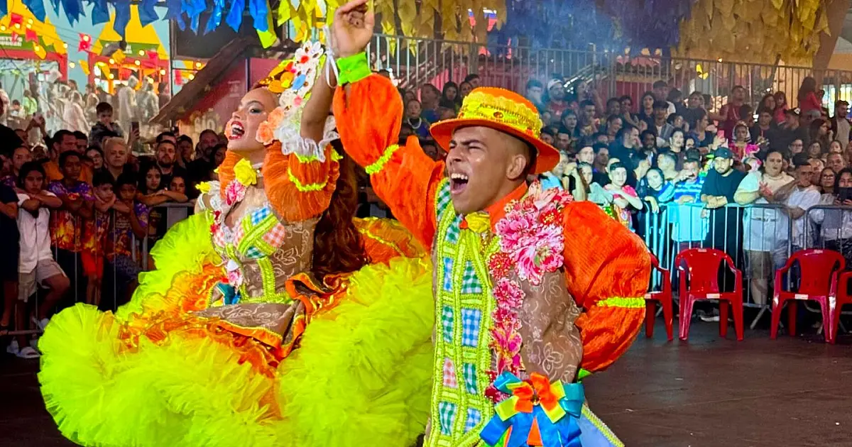 Casais vestidos com trajes típicos juninos dançando alegremente em um grande pavilhão decorado com bandeirinhas coloridas