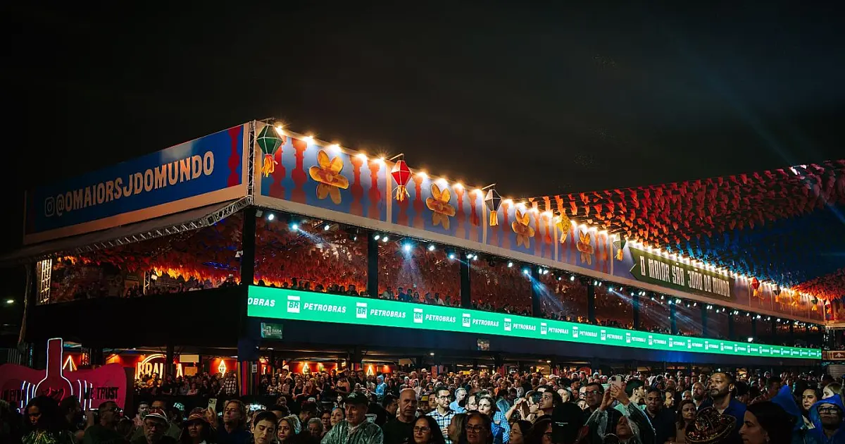 Imagem panorâmica noturna com plano aberto mostrando o Parque do Povo completamente lotado de pessoas com os braços levantados em direção ao palco iluminado e decorado com bandeirinhas coloridas