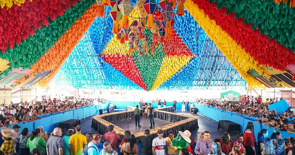 Imagem aérea de uma multidão vestida com trajes xadrez coloridos dançando em uma grande praça pública enfeitada com bandeirinhas