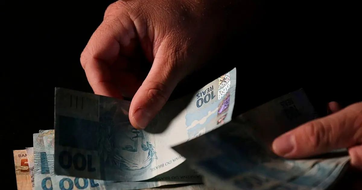 Imagem em close-up focando nas mãos de uma pessoa manuseando e contando diversas cédulas de cem reais sobre uma superfície lisa