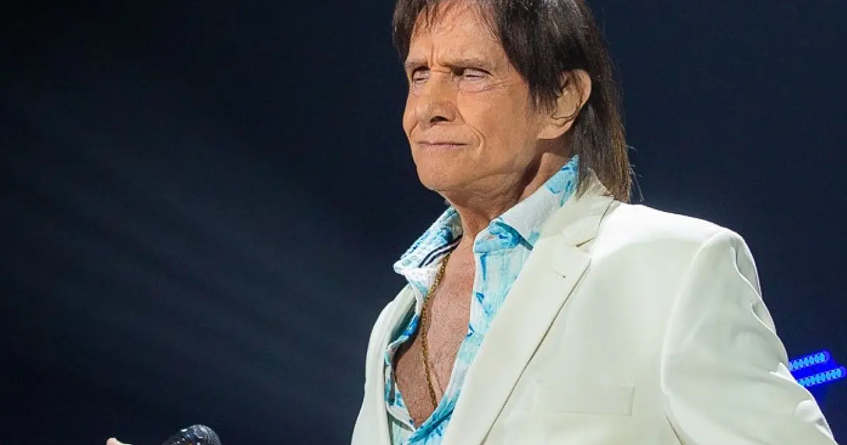 O cantor Roberto Carlos vestindo blazer azul e sorrindo durante uma apresentação musical iluminada no palco