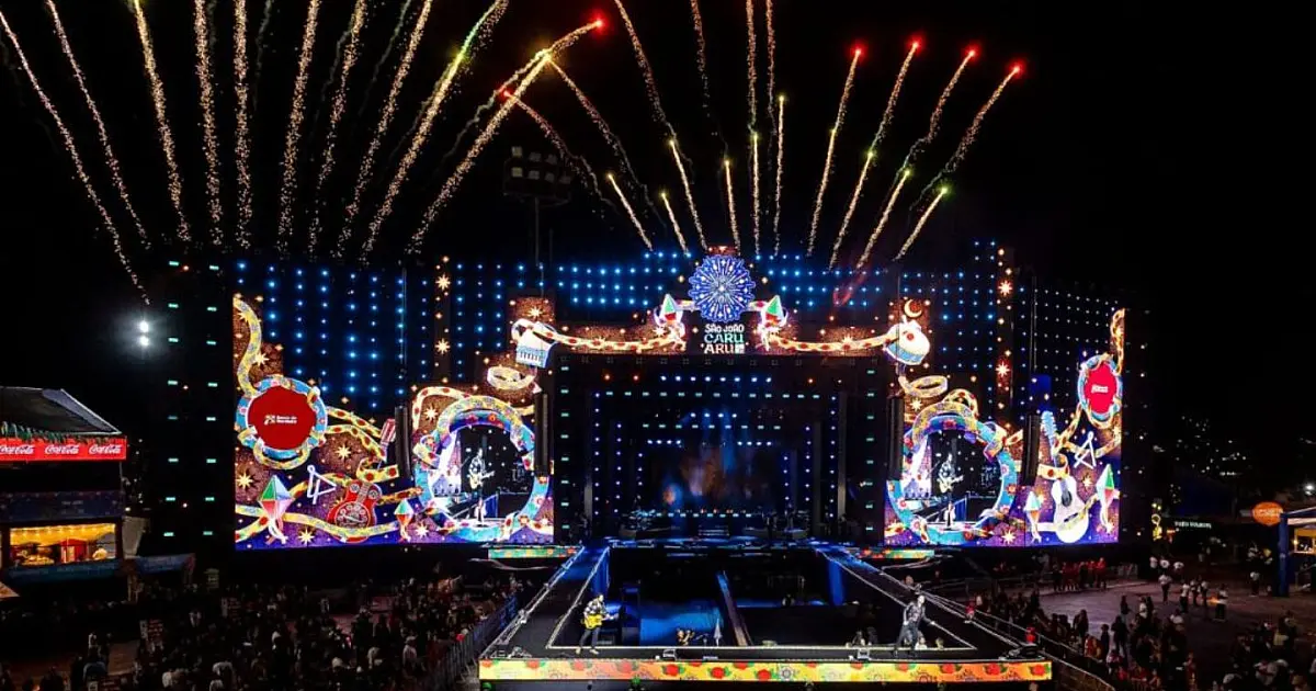 Vista aérea panorâmica e noturna do Pátio de Eventos em Caruaru lotado de pessoas, com o palco principal iluminado ao fundo e bandeirinhas juninas estendidas sobre a multidão