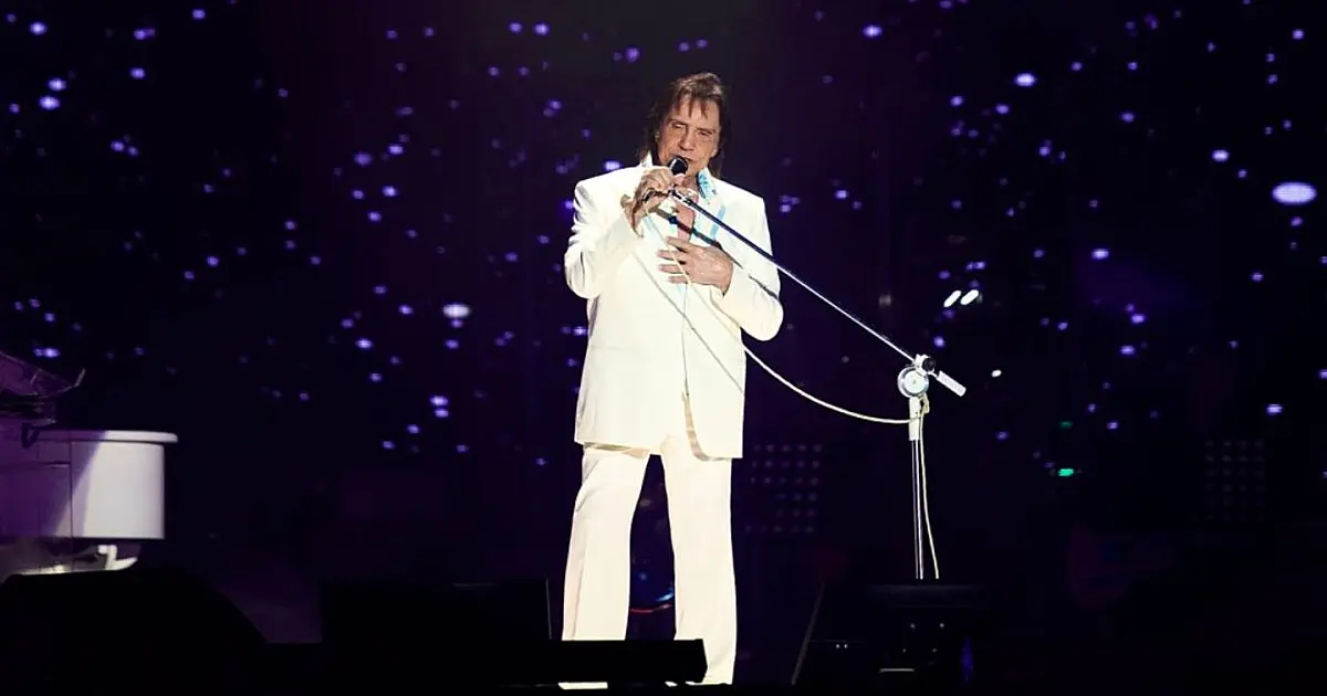 O cantor Roberto Carlos vestindo seu clássico terno azul e branco, cantando com um microfone na mão sob as luzes de um grande palco iluminado.