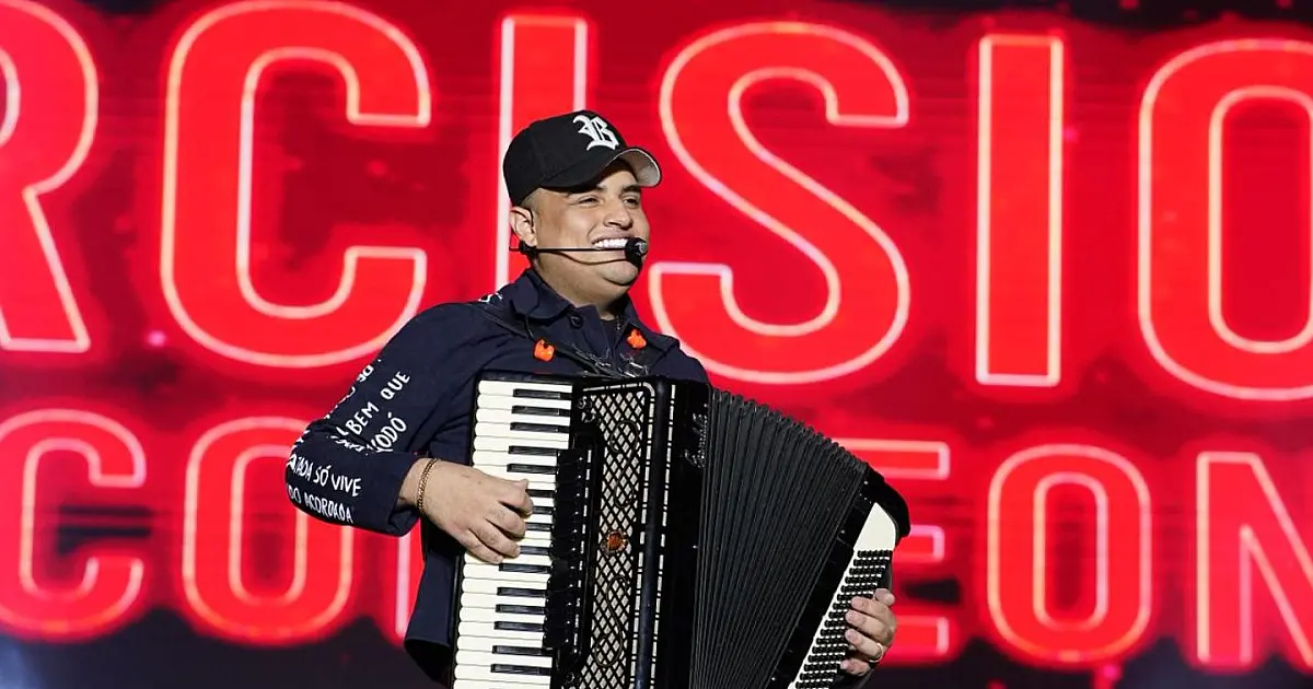 Cantor no palco principal segurando um microfone e apontando para uma plateia iluminada e gigantesca durante a noite