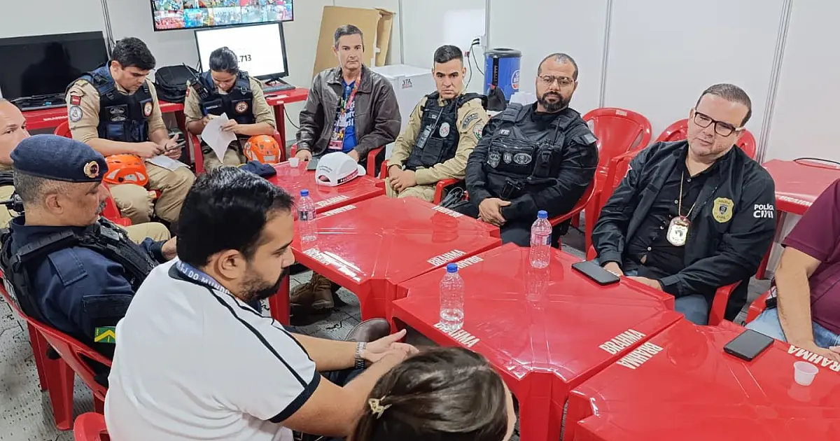 Profissionais de colete azul da assistência social conversando com policiais militares fardados dentro de uma sala de comando montada com telas e computadores