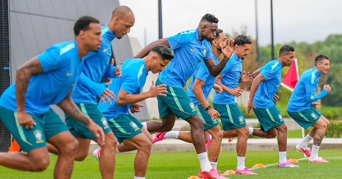 Grupo de jogadores da Seleção Brasileira perfilados no gramado verde durante atividade tática, ouvindo orientações de Carlo Ancelotti no centro do campo