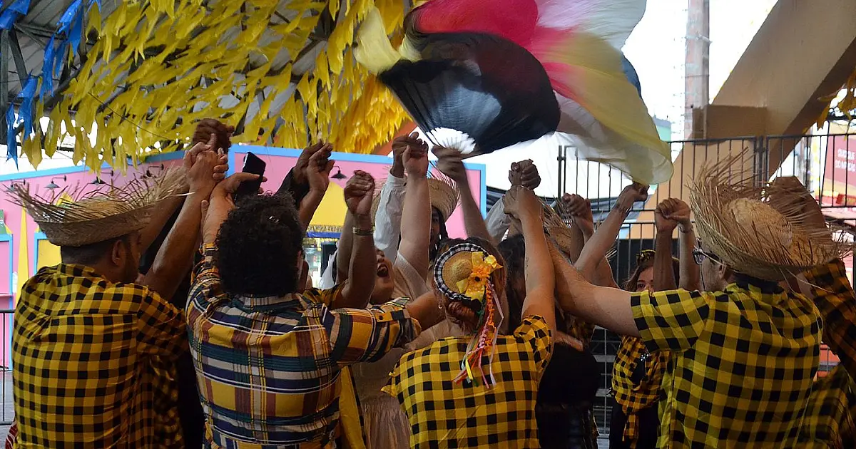 Grupo de pessoas vestidas com trajes juninos coloridos dançando e sorrindo em círculo dentro de uma estrutura em formato de pirâmide decorada com bandeirolas.