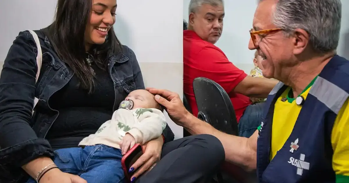 Profissional de saúde de luvas brancas aplicando uma injeção com seringa no braço de uma criança pequena sentada no colo da mãe em uma sala de atendimento de uma UBS