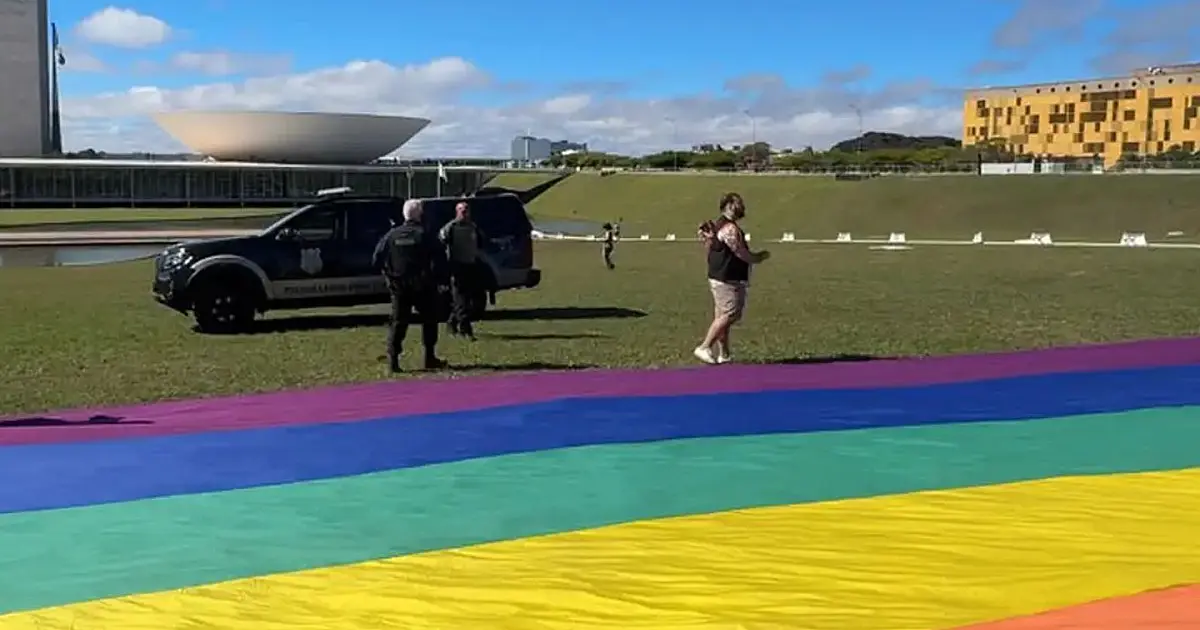 Vista ampla do gramado em frente ao Congresso Nacional, onde um grupo de pessoas começa a desdobrar uma longa e extensa bandeira com as cores do arco-íris sob o céu aberto de Brasília