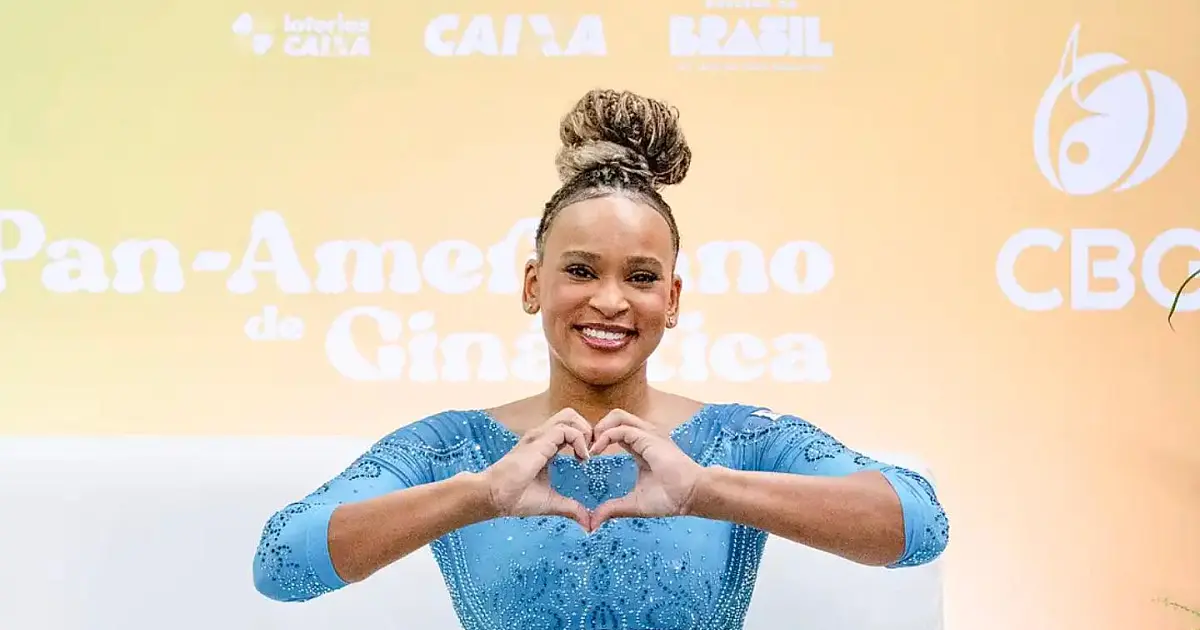 A ginasta Rebeca Andrade sorrindo no topo do pódio vestindo o uniforme oficial do Brasil e segurando a medalha de ouro no peito