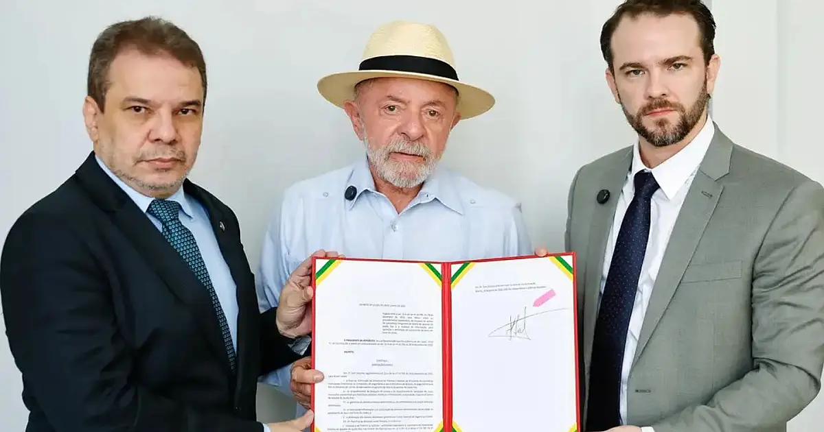 O presidente Luiz Inácio Lula da Silva sentado à mesa presidencial em seu gabinete, assinando um documento oficial com uma caneta sob a luz de luminárias de escritório
