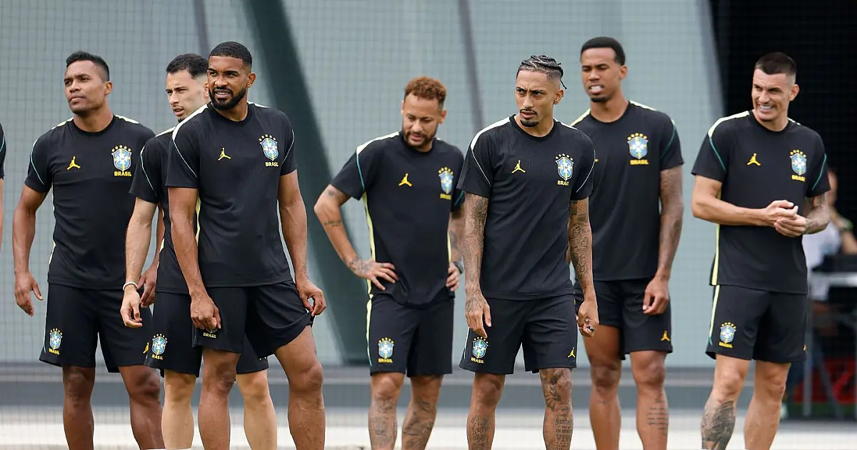 Jogadores com o colete azul de treino da Seleção Brasileira correndo pelo gramado verde em um dia ensolarado