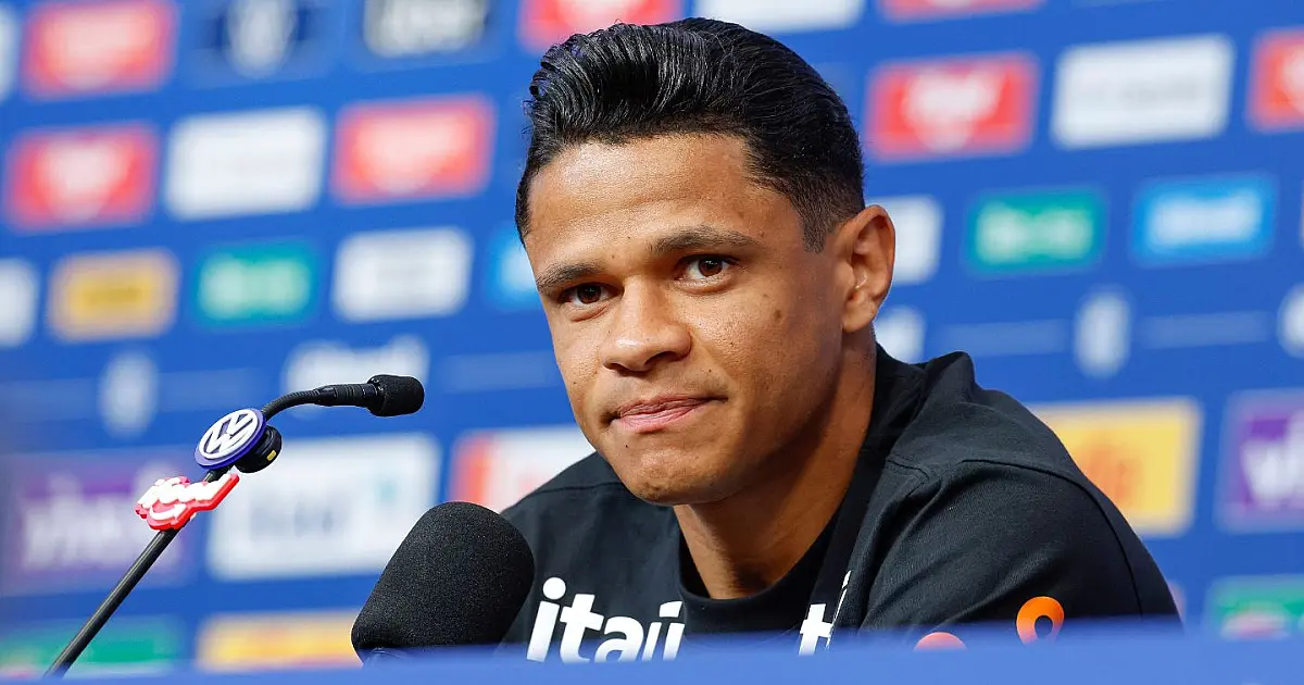 Jogador de futebol sorridente usando agasalho oficial de treino azul marinho, sentado em uma mesa de coletiva repleta de microfones