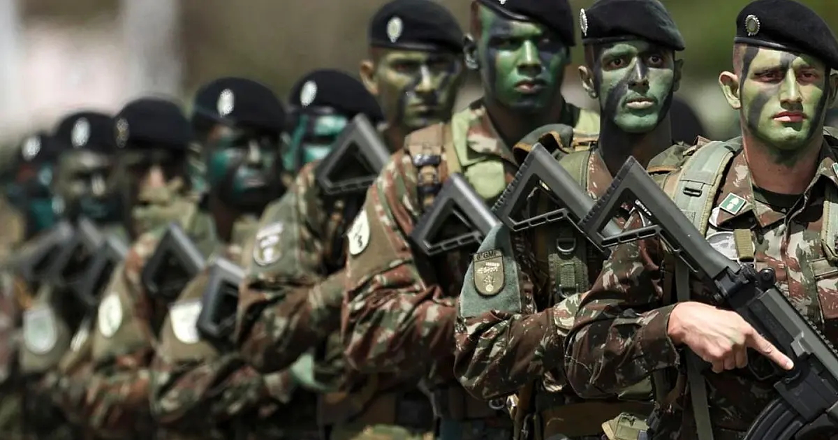 Soldados do Exército Brasileiro fardados marchando em formação com equipamentos de segurança em uma rua asfaltada durante o dia