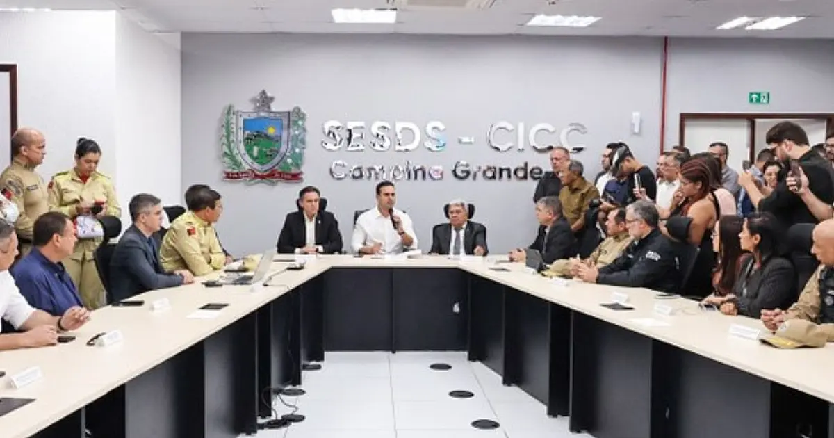 Autoridades públicas reunidas em uma sala de monitoramento com grandes telas exibindo mapas e gráficos estatísticos