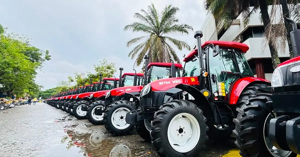 Fileira de microtratores vermelhos e equipamentos agrícolas alinhados em um pátio aberto, com autoridades públicas ao fundo conversando durante evento oficial sob luz do dia
