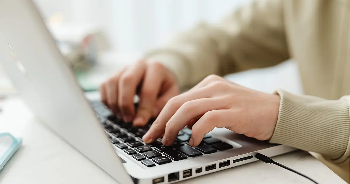 Mãos de um profissional digitando no teclado de um notebook moderno em cima de uma mesa escura sob iluminação de escritório